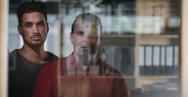 Zwei Männer stehen hintereinander vor der spiegelnden Scheibe eines Fensters