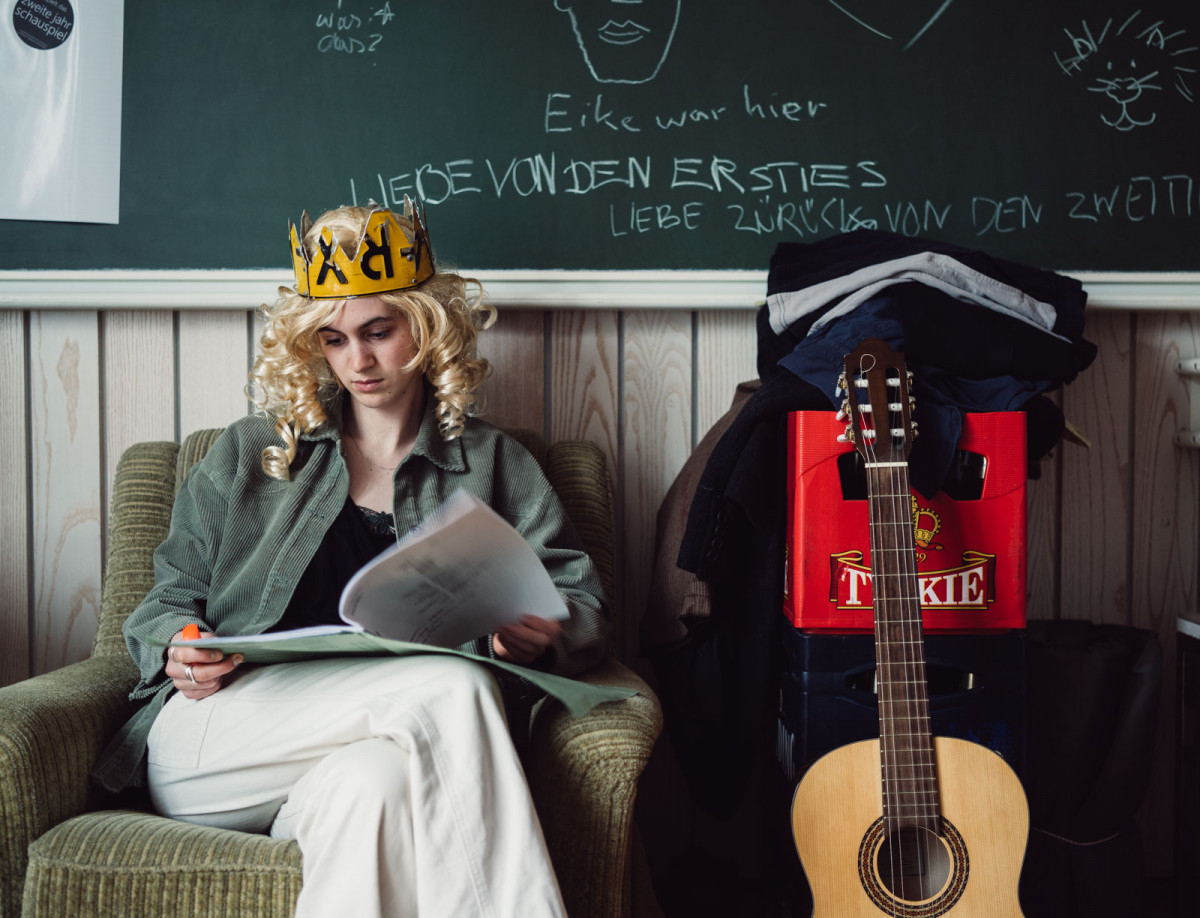 Schauspielstudentin Fenna sitzt mit blonder Perrücke und einer Krone in einem Cordsessel und geht einen Text durch.