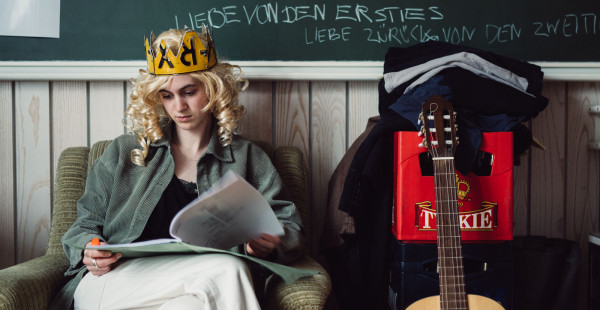 Schauspielstudentin Fenna sitzt mit blonder Perrücke und einer Krone in einem Cordsessel und geht einen Text durch.