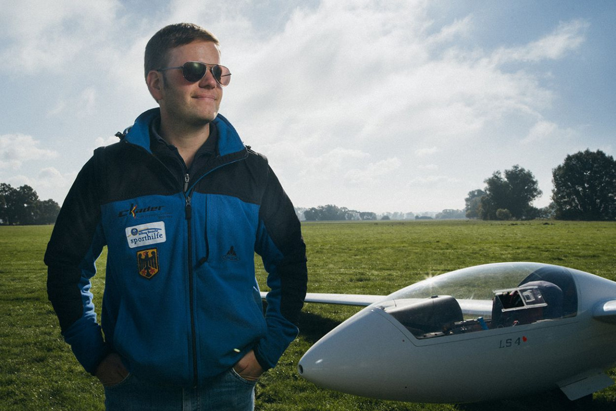 Ein junger Mann mit Sonnenbrille steht zufrieden neben einem Segelflugzeug