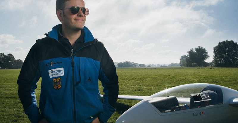 Ein junger Mann mit Sonnenbrille steht zufrieden neben einem Segelflugzeug