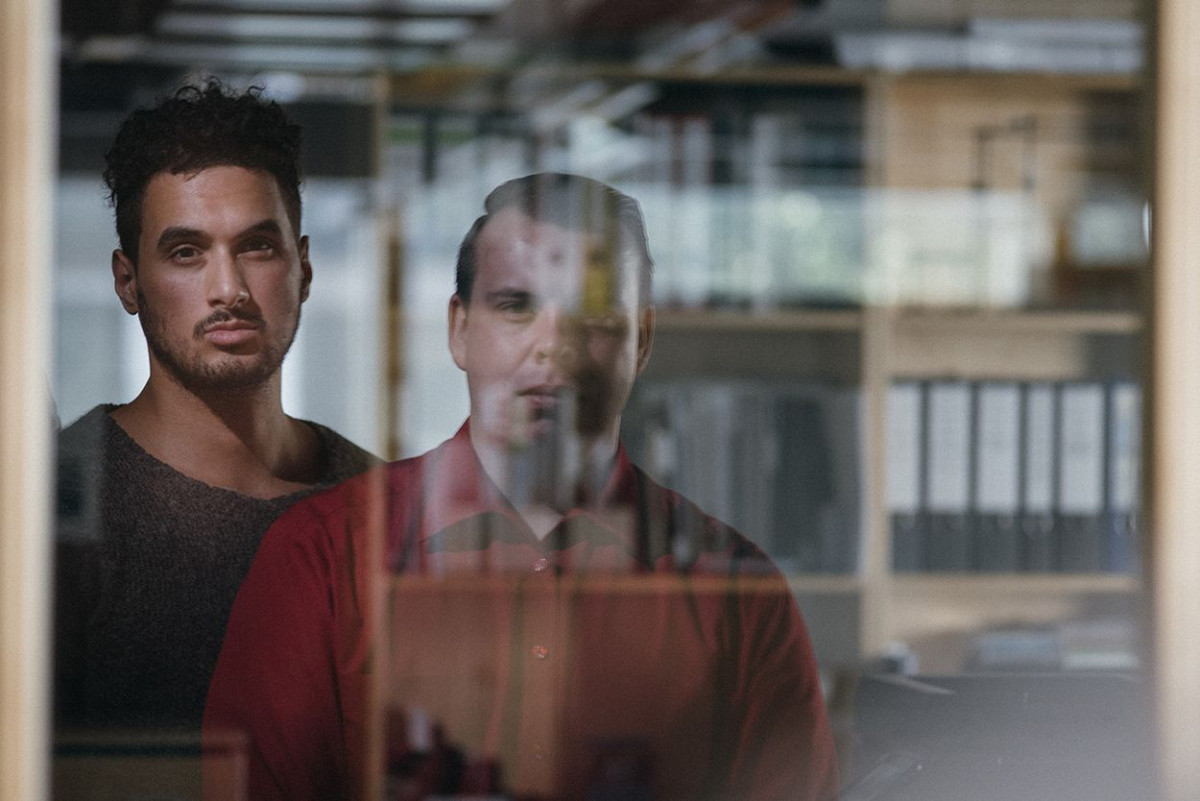 Zwei Männer stehen hintereinander vor der spiegelnden Scheibe eines Fensters