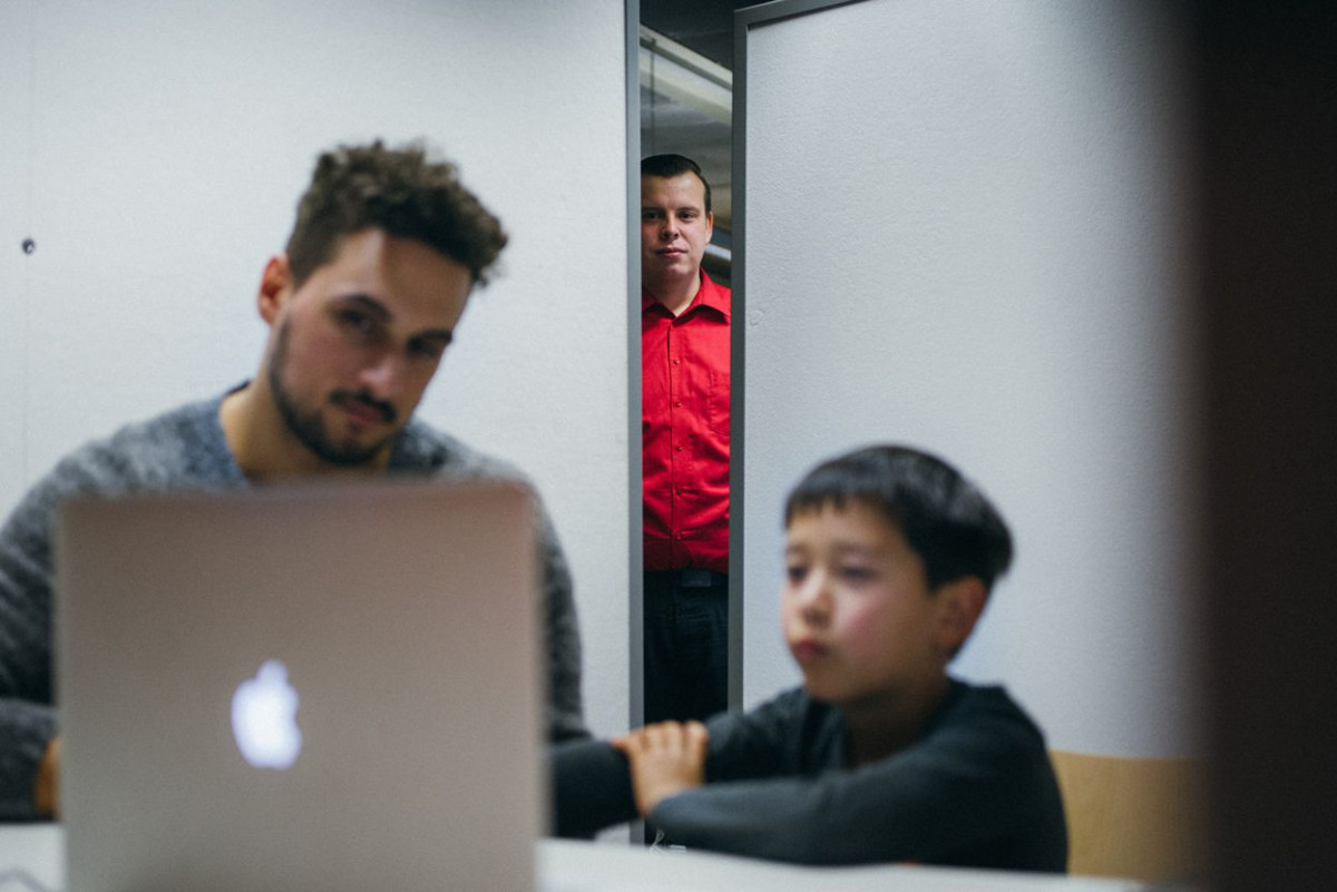 Ein Mann schaut zusammen mit einem jungen auf einen Laptop, während ein anderer Mann durch einen Türspalt zusieht